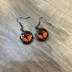 May include: A pair of silver-toned dangle earrings. Each earring features a circular orange center with a black design, surrounded by a silver frame. The earrings are displayed on a wooden surface.