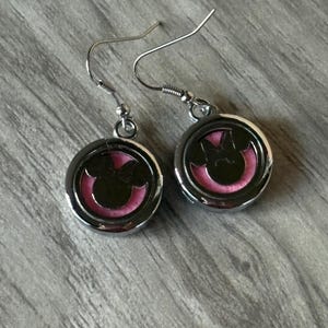 May include: A pair of silver-toned dangle earrings. Each earring features a circular charm with a black silhouette of a bow and mouse ears, set against a pink background. The earrings have a simple hook design.