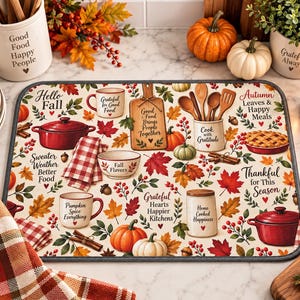 May include: A rectangular kitchen drying mat featuring a cream background with a pattern of red Dutch ovens, pumpkins, autumn leaves, berries, and kitchenware. Text includes Hello Fall, Good Food Brings People Together, and Pumpkin Spice Everything.