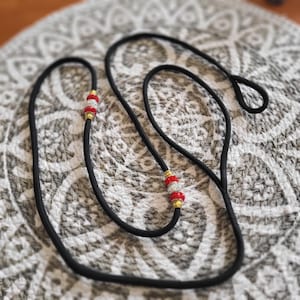 May include: Black rope leash with red and white beaded accents and gold-colored metal details. The leash is looped at one end. The background is a gray and white patterned surface.