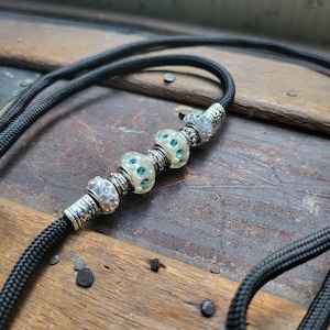 May include: Black nylon cord with decorative beads. The beads are a mix of iridescent and light blue, with silver-toned metal accents. The cord is thick and appears durable, suitable for a necklace or similar accessory.