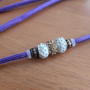 May include: Close-up of a purple cord with decorative beads. The beads are a mix of silver, gold, and clear crystal-like accents. The cord has a braided texture, and the beads are clustered together, creating a decorative element.
