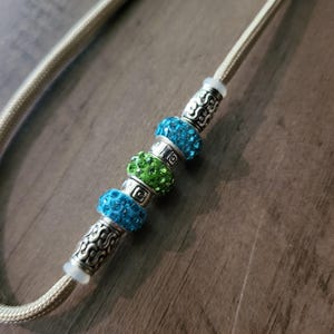 May include: A close-up of a beaded necklace with a beige cord. The necklace features silver-colored metal accents and sparkling beads in shades of blue and green. The beads are interspersed with decorative metal spacers.
