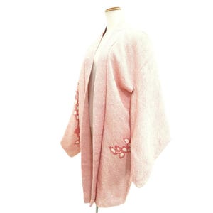 May include: A light pink haori jacket featuring a textured fabric surface and a subtle white floral pattern on the sleeves and lower front panels. The garment has wide, loose sleeves, an open front design, and a straight, structured silhouette.