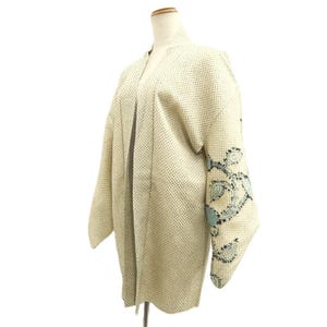 May include: A cream-colored haori jacket featuring a textured, grid-like pattern across the fabric. The garment has wide sleeves adorned with a decorative, swirling floral embroidery in shades of light blue and black along the lower edges.