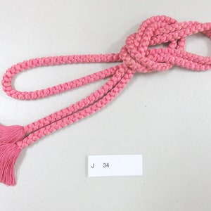 Könnte beinhalten: Eine rosa geflochtene Schnur mit Quasten an jedem Ende. Die Schnur ist zu einem Knoten gebunden und das Etikett "J 34" ist sichtbar.