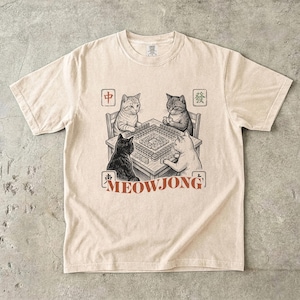 May include: Beige t-shirt featuring a detailed illustration of four cats playing mahjong. The word "MEOWJONG" is printed in red below the table. The cats are depicted in a vintage, hand-drawn style.
