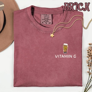 May include: Brick-colored t-shirt with a crew neck and short sleeves. The shirt features an embroidered beer glass design with the text "VITAMIN G" below it. A gold necklace with a star pendant is draped across the shirt.