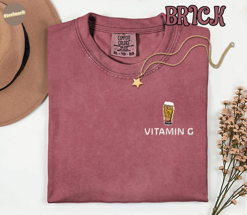 May include: Brick-colored t-shirt with a crew neck and short sleeves. The shirt features an embroidered beer glass design with the text "VITAMIN G" below it. A gold necklace with a star pendant is draped across the shirt.