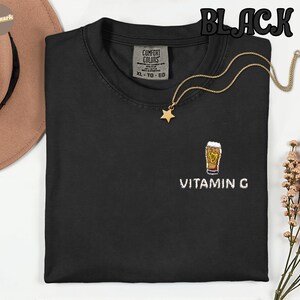 May include: Black crew neck t-shirt with an embroidered beer glass design and the text "VITAMIN G". A gold necklace with a star pendant is draped on the shirt. The word "BLACK" is printed at the top.