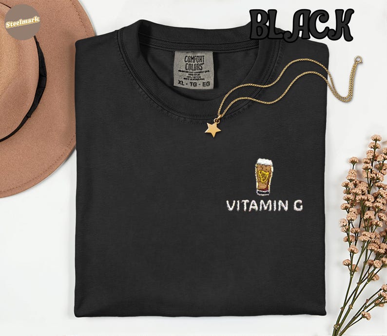 May include: Black crew neck t-shirt with an embroidered beer glass design and the text "VITAMIN G". A gold necklace with a star pendant is draped on the shirt. The word "BLACK" is printed at the top.