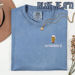 May include: A light blue Comfort Colors t-shirt with the text "BLUE JEAN" at the top. The shirt has an embroidered design of a beer glass with the text "VITAMIN G" below it. A gold star pendant necklace is draped on the shirt.