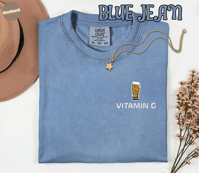 May include: A light blue Comfort Colors t-shirt with the text "BLUE JEAN" at the top. The shirt has an embroidered design of a beer glass with the text "VITAMIN G" below it. A gold star pendant necklace is draped on the shirt.