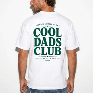 May include: White t-shirt with the text "COOL DADS CLUB" in dark green, along with "PREMIUM MEMBER OF THE" above and "POWERED BY LOVE & ESPRESSO EST.2026" below. The shirt is a casual wear item.