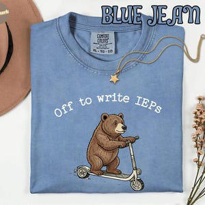 Könnte beinhalten: Blaues Jeans-T-Shirt mit dem Text "Off to write IEPs" und einem Cartoon-Bären auf einem Roller. Das Shirt hat einen Rundhalsausschnitt und kurze Ärmel. Eine goldene Stern-Anhänger-Halskette ist über dem Shirt drapiert.