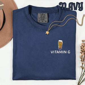May include: Navy blue t-shirt with a small embroidered beer glass design and the text "VITAMIN G". The shirt has a crew neck and short sleeves. A gold-colored necklace with a star pendant is draped on the shirt.