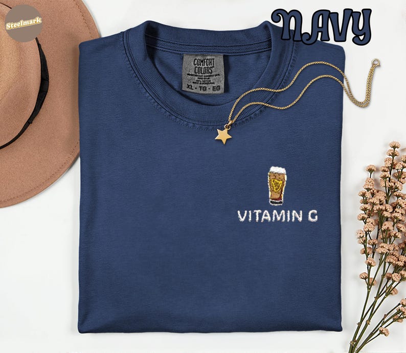 May include: Navy blue t-shirt with a small embroidered beer glass design and the text "VITAMIN G". The shirt has a crew neck and short sleeves. A gold-colored necklace with a star pendant is draped on the shirt.