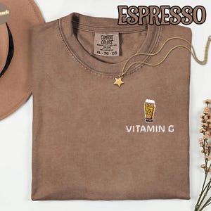 May include: Espresso-colored Comfort Colors t-shirt with a crew neck and short sleeves. The shirt features an embroidered design of a beer glass with the text "VITAMIN G" below it. A gold necklace with a star pendant is draped on the shirt.