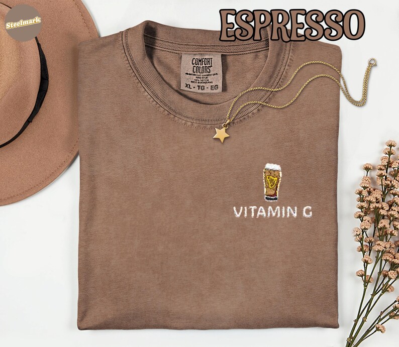 May include: Espresso-colored Comfort Colors t-shirt with a crew neck and short sleeves. The shirt features an embroidered design of a beer glass with the text "VITAMIN G" below it. A gold necklace with a star pendant is draped on the shirt.