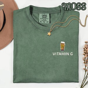 May include: A moss green XL Comfort Colors t-shirt with an embroidered beer glass design and the text "VITAMIN G". A gold star pendant necklace rests on the shirt. The word "MOSS" is in a green, stylized font.