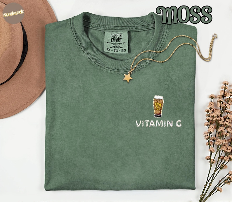 May include: A moss green XL Comfort Colors t-shirt with an embroidered beer glass design and the text "VITAMIN G". A gold star pendant necklace rests on the shirt. The word "MOSS" is in a green, stylized font.