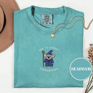 May include: Seafoam green t-shirt with a cartoon koala wearing a wizard robe and holding a staff. The text "OFF TO COMMIT SHENANIGANS" is above and below the koala. A gold star pendant necklace is on the shirt.