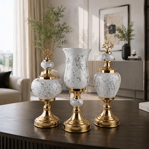 Puede incluir: Un conjunto de tres recipientes decorativos con un patrón de mármol blanco y negro, bases de metal dorado pulido y detalles. Dos recipientes tienen tapas con remates dorados en forma de coral, mientras que el central es un jarrón alto.