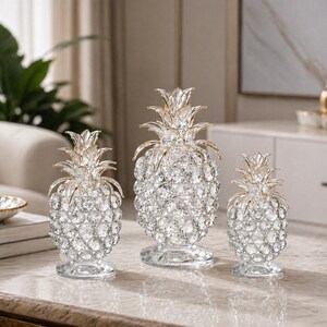Puede incluir: Tres esculturas de piña de cristal de diferentes tamaños. Cada piña tiene una corona dorada y está cubierta de cristales transparentes y brillantes. Las esculturas se asientan sobre una base redonda transparente, añadiendo un toque de elegancia a cualquier decoración.