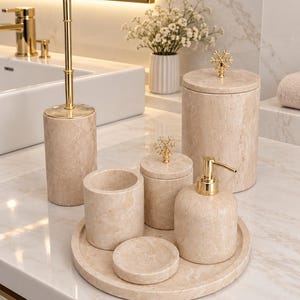 Könnte beinhalten: Ein Badezimmer-Zubehörset aus beigem Marmor, bestehend aus einem Seifenspender, einem Zahnbürstenhalter, einer Seifenschale, einem Behälter mit Deckel und einem Toilettenbürstenhalter. Jedes Teil hat goldfarbene Metallakzente und schneeflockenförmige Knäufe auf den Deckeln.
