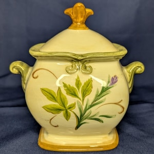 Op de afbeelding: Een crèmekleurige keramische suikerpot met bijpassend deksel, voorzien van een handgeschilderd ontwerp van groene bladeren en paarse bloemen. De pot heeft twee zijhandvatten, een vierkante voet en een gele knop, alles met craquelé glazuur.