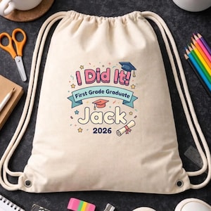 May include: A cream-colored drawstring backpack with the text "I Did It! First Grade Graduate Jack 2026" in pink, blue, and yellow. The design includes a graduation cap, stars, and a diploma. The bag has white drawstrings.