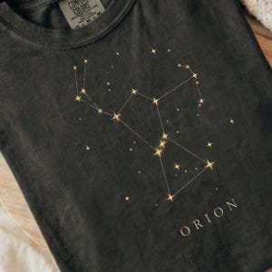 May include: A charcoal gray t-shirt featuring a gold-colored graphic of the Orion constellation with connected lines and star points. The word ORION is printed in gold capital letters below the constellation. The shirt has a Comfort Colors label.