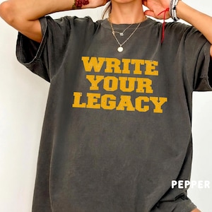 May include: A charcoal gray short-sleeve t-shirt featuring the text "WRITE YOUR LEGACY" in a distressed, bold yellow font across the chest. The garment has a crew neckline and a relaxed fit, with the word "PEPPER" printed in small white text.
