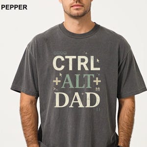 May include: Dark gray t-shirt with the text "CTRL + ALT + DAD" in white and green. The shirt has a vintage, faded look. The word "PEPPER" is printed in the upper left corner.