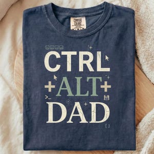 May include: Navy blue t-shirt with the text "CTRL + ALT + DAD" in a distressed font. The design includes computer-related symbols. The shirt is folded on a wooden surface.