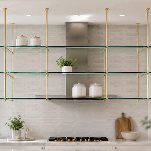 Op de afbeelding: Een hangend rek met drie niveaus, bestaande uit heldere glazen planken ondersteund door een messingkleurig metalen frame. De constructie is aan het plafond bevestigd en bevat witte keramische potten en een kleine groene plant.