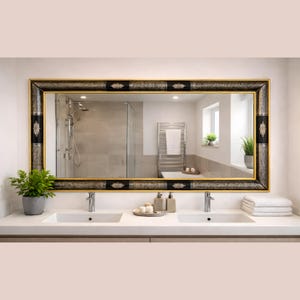 May include: Rectangular bathroom mirror with a black and gold ornate frame. The mirror reflects a modern bathroom with a shower, towel rack, and double sink vanity. The frame has decorative black panels and gold accents.