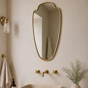 Könnte beinhalten: Ein goldgerahmter Spiegel mit gewelltem Rand hängt über einem Waschbecken im Badezimmer. Der Spiegel spiegelt einen Blick in den Raum wider. Ein goldener Wasserhahn und passende Armaturen sind an der Wand angebracht. Eine Vase mit Grün steht auf der Theke.
