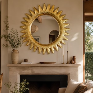 May include: A large, circular sunburst mirror with a textured, metallic gold frame featuring radiating spikes. The mirror has a reflective glass center and is mounted on a wall above a stone fireplace mantel, reflecting a room interior.
