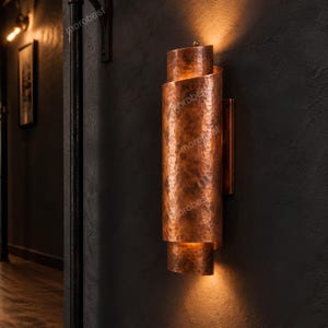 May include: A wall sconce featuring a cylindrical, hammered copper finish with overlapping metal panels. The fixture emits a warm, diffused light from the top and bottom openings, casting a glow against the dark wall surface behind it.