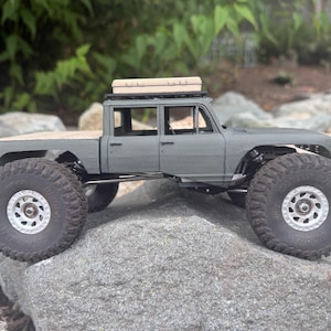May include: A matte gray scale model pickup truck with a four-door cab, a flatbed, and large black off-road tires with silver rims. A tan rectangular storage case is mounted on the roof rack of the vehicle, which is positioned on a rock.