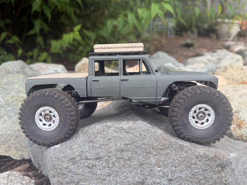 May include: A matte gray scale model pickup truck with a four-door cab, a flatbed, and large black off-road tires with silver rims. A tan rectangular storage case is mounted on the roof rack of the vehicle, which is positioned on a rock.