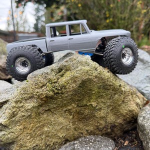 May include: A gray radio-controlled truck model with a crew cab body style, featuring large black off-road tires with deep treads, silver rims, and a visible suspension system with blue accents, positioned on a large, textured rock.