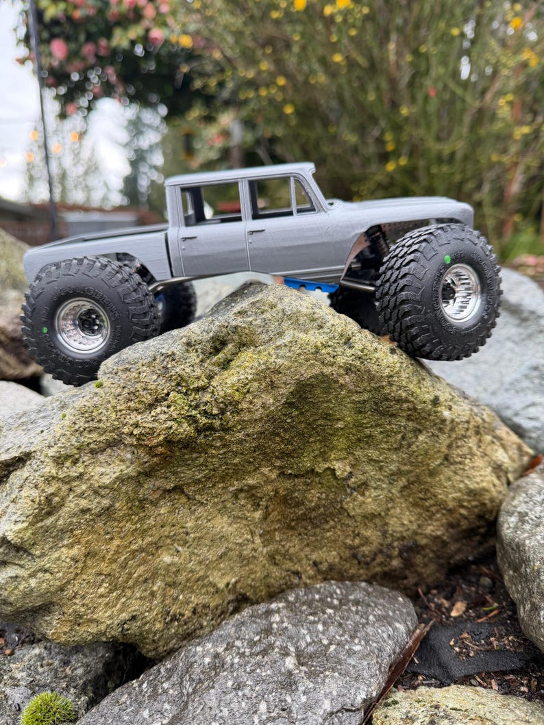 May include: A gray radio-controlled truck model with a crew cab body style, featuring large black off-road tires with deep treads, silver rims, and a visible suspension system with blue accents, positioned on a large, textured rock.