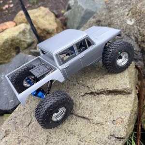 May include: A silver remote-controlled pickup truck model featuring large black off-road tires, metallic silver wheels, and visible blue suspension components. The truck has an open cargo bed and a detailed cabin interior visible through the windows.