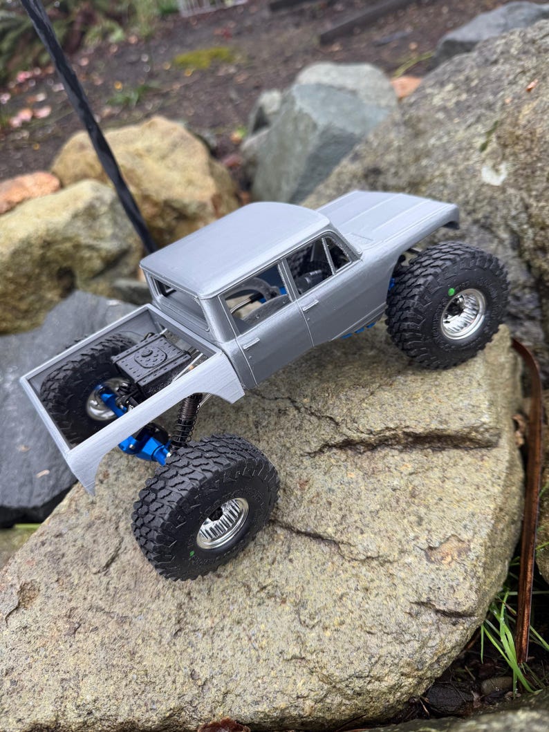 May include: A silver remote-controlled pickup truck model featuring large black off-road tires, metallic silver wheels, and visible blue suspension components. The truck has an open cargo bed and a detailed cabin interior visible through the windows.