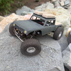 May include: A matte olive green remote-controlled crawler truck featuring large black off-road tires, silver wheels, and two tan storage cases mounted on a black roof rack. The vehicle has yellow circular headlights and an open chassis.