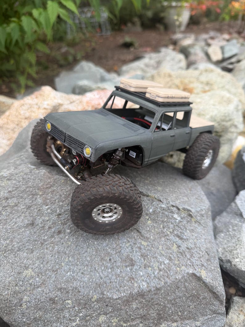 May include: A matte olive green remote-controlled crawler truck featuring large black off-road tires, silver wheels, and two tan storage cases mounted on a black roof rack. The vehicle has yellow circular headlights and an open chassis.