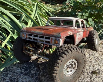 May include: A remote-controlled truck with a weathered, rust-colored body and a white roof. The vehicle features large, knobby black tires with silver rims, a white front grille, and circular yellow KC-branded headlights on the front bumper.