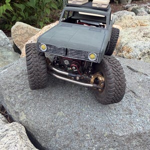 May include: A gray remote-controlled crawler truck featuring large treaded tires, a black roof rack with two beige storage cases, yellow circular headlights, and a visible red winch component mounted on the front bumper assembly.